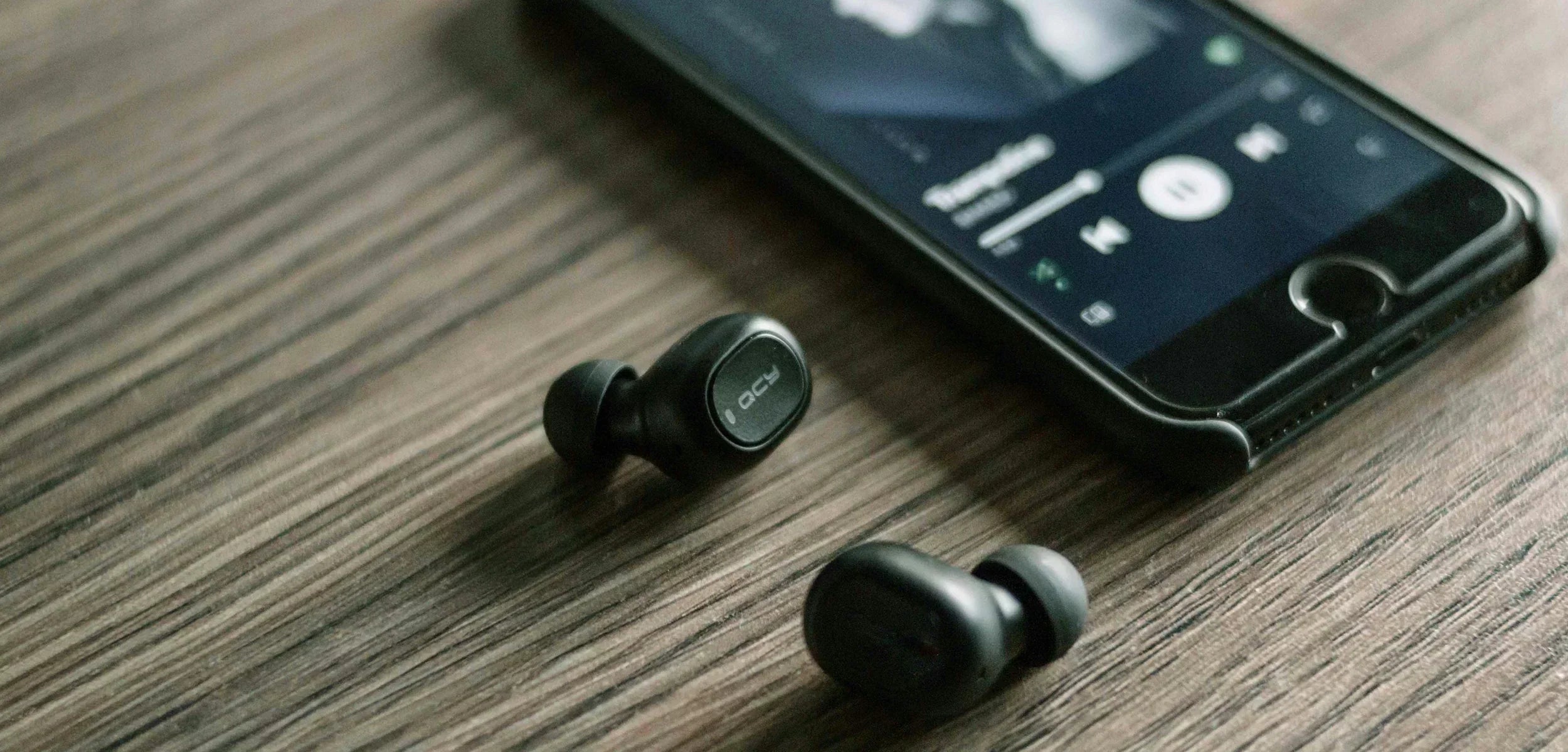 Schwarze QCY T1 Bluetooth-Kopfhörer neben Smartphone auf Holzoberfläche.