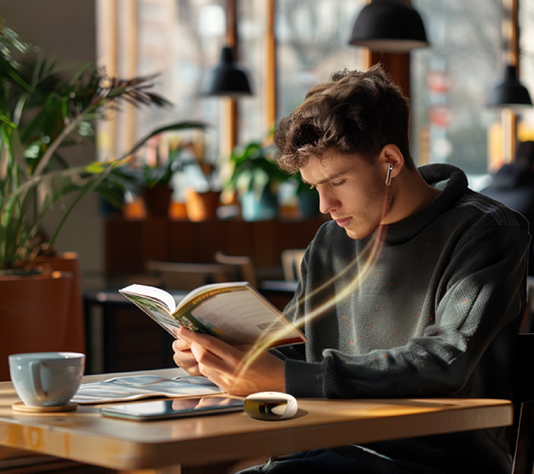 Giovane uomo legge un libro in un caffè con cuffie wireless e tazza di caffè sul tavolo di legno.