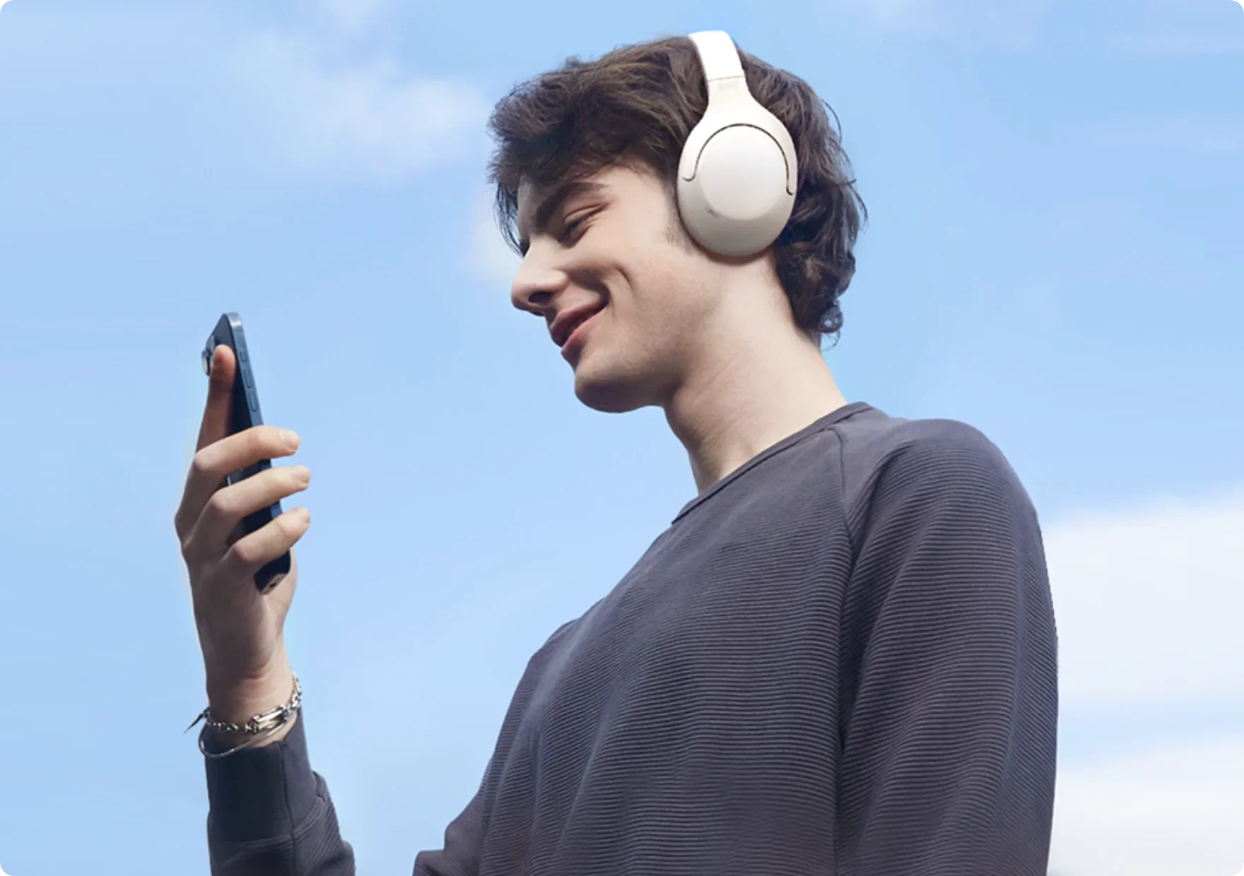 Giovane uomo con cuffie bianche e smartphone sotto un cielo azzurro.