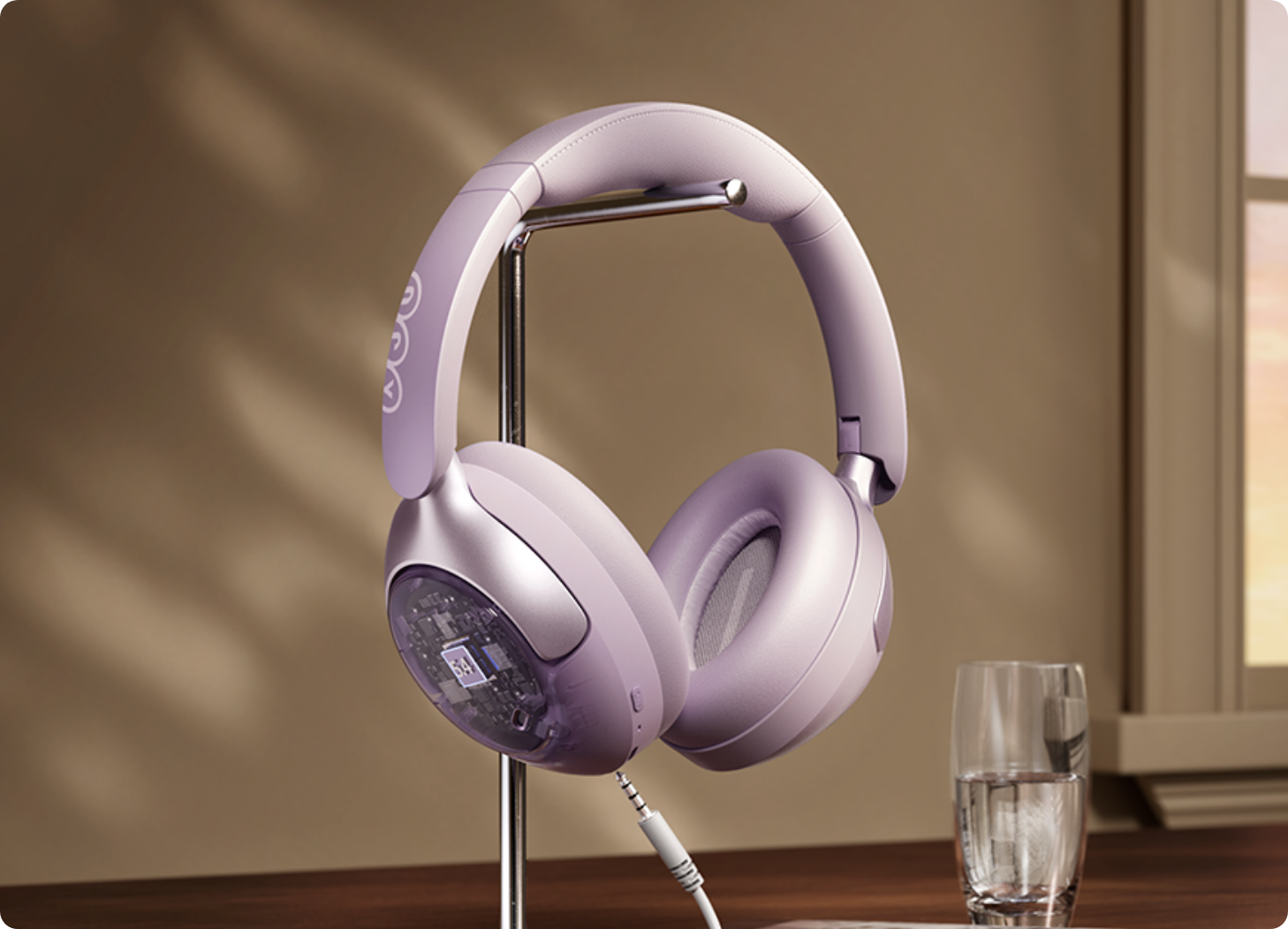Cuffie over-ear color lavanda con custodia trasparente su supporto, cavo collegato, accanto a un bicchiere d'acqua sul davanzale della finestra.